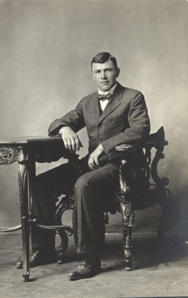 Full-length seated studio portrait of John Maly, wearing a suit and bow tie. He is seated in a chair with his right forearm resting on a table.
