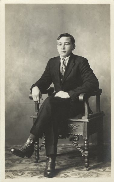 Full-length seated studio portrait of Edwin Schumann wearing a suit in front of a painted backdrop. His legs are crossed, resting one hand on his leg, the other on the arm of the chair.