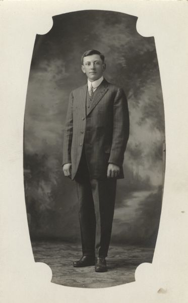 Full-length studio portrait of Charles Zander standing in front of a painted backdrop wearing a suit. The portrait has a white, decorative border.
