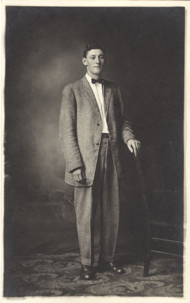 Full-length studio portrait of Max Esser standing and wearing a suit and bow tie in front of a painted backdrop. His hand rests on the back of a chair.