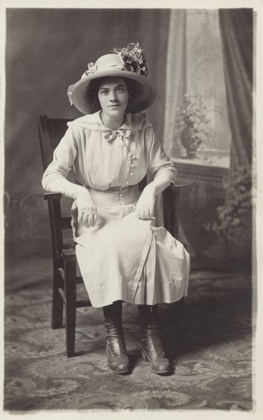Full-length studio portrait of Lena Gerath Falkenstein seated in a chair on a patterned carpet in front of a painted backdrop. She is wearing a dress, flowered hat, button shoes and white gloves.