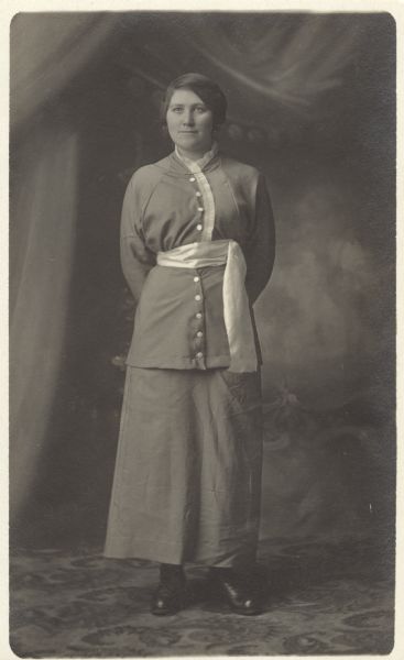Full-length studio portrait of Nellie Faust standing on a patterned carpet in front of a painted background. She is wearing a skirt and tunic with a scarf tied as a belt. Her hands are clasped behind her back.