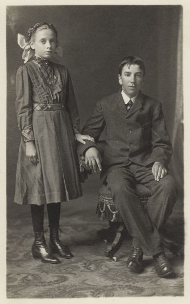 Full-length studio portrait of Elizabeth and Clemens Hellebrand in front of a painted backdrop. He is sitting in a chair, and she is standing and resting her hand on his sleeve. They may be brother and sister. She is wearing a dress and large hair bow. He is wearing a suit.