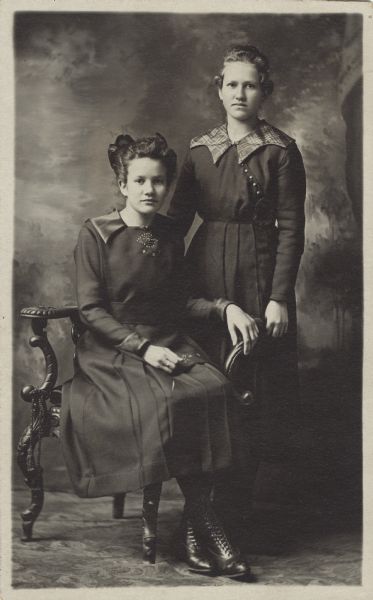 Full-length studio portrait of Mary and Elizabeth Holfelder in front of a painted backdrop. They are possibly sisters. Both are wearing dresses, and the woman sitting has a hair bow.