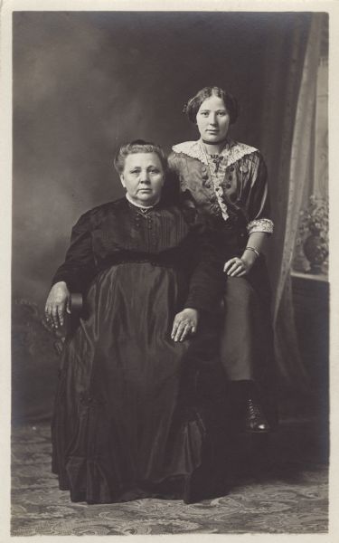 Full-length studio portrait of two women, one seated and the other perched on the chair arm, posed in front of a painted backdrop. They are wearing dresses. Three names are written on the back of the postcard; Mrs. Matt Annen, Mrs. Allie Statz and Connie Annen.