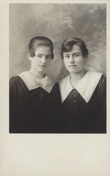 Quarter-length studio portrait of Mary Gorman and Amanda Faust in front of a painted backdrop. They are wearing dark dresses with light collars.