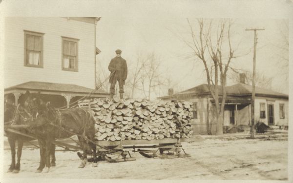 A man is standing on top of a stack of firewood that is loaded on his horse-drawn bobsled. He is wearing a coat and hat. In the background are two homes and several trees.