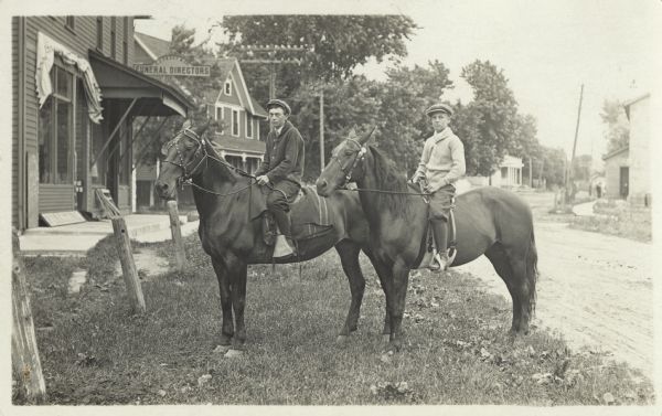 Two young men are posing outdoors on horseback in front of the C.J. Faust & Co., Funeral Directors' building. They are wearing jackets and hats. A dirt road is behind them and buildings are on each side.