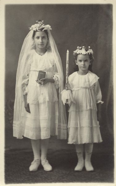 Photographic postcard of a full-length studio portrait of two unidentified girls on the occasion of their First Communion. They are standing and wearing white dresses and floral head wreaths. The girl on the left is wearing a veil and holding a catechism and rosary in her left hand; the girl on the right is holding a decorative candle in her right hand. They are posing in front of a painted backdrop.