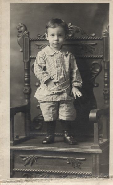 Full-length studio portrait of a young boy, standing on the seat of an ornately carved chair. He is wearing a checkered outfit with stockings and shoes. His right hand is resting inside his belt.