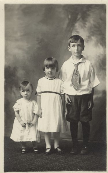 Full-length studio portrait of two boys and a girl in front of a painted backdrop. The girl and younger boy are wearing dresses, white stockings and dark shoes. The older boy is wearing knickers (knickerbockers), a shirt, tie, dark stockings and shoes. Names, (l to r), are Melchoir Coyle (brother), Mary Coyle (sister) and Mathew J. Coyle (brother). All are children of Joseph Coyle and Katherine Endres Coyle.