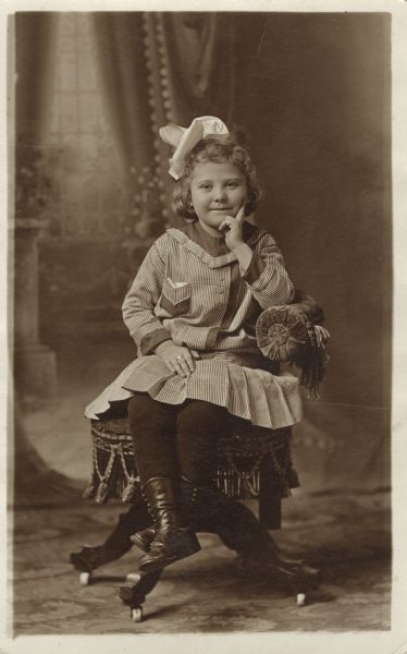 Full-length studio portrait of a girl, seated in a chair with fringe, in front of a painted backdrop. She has her left hand resting on her chin and is wearing a dress, large hair bow, stockings and shoes.