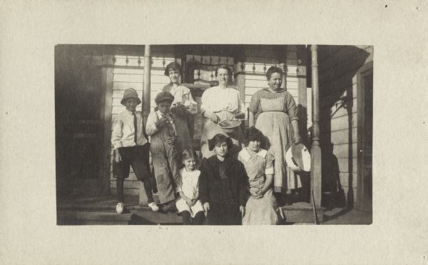Group portrait of a family posed on a porch. The three women in the back hold a large enamel bowl, a mixing bowl and spoon, and a grinder or sifter. In front are two young boys and three girls.