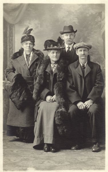 Full-length studio portrait of two men and two women in front of a painted backdrop. Two are seated, the other two are standing. They are dressed in winter coats and hats, the women's hats decorated with feathers. The women have fur collars, stoles and muffs.