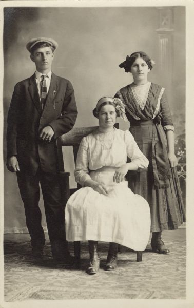 Full-length studio portrait of two women and one man in front of a painted backdrop. One women is seated in a wooden chair, the others are standing on each side. Both women wear ribbons in their hair.
