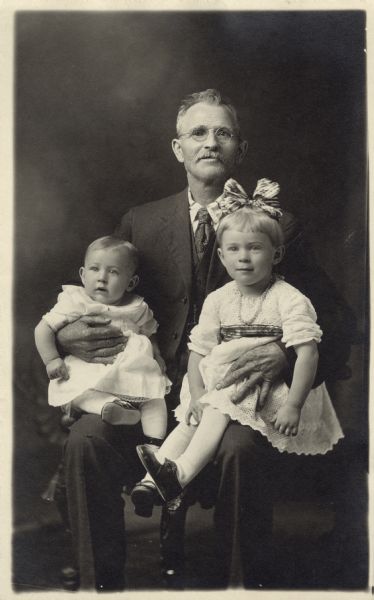 Full-length studio portrait of a seated man with a boy and a girl, one on each knee. They are posed in front of a painted backdrop.