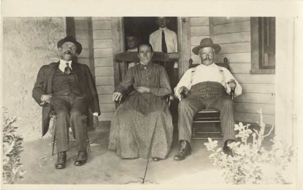 Two men and one woman are seated on a porch. The men are smoking cigars. Two children can be seen standing behind the screen door behind them.
