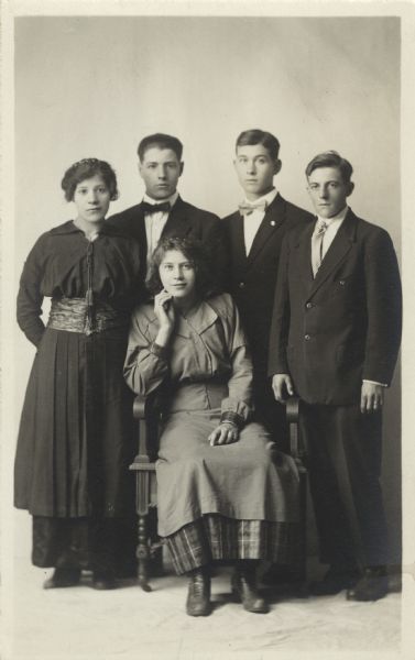 Full-length studio portrait of five teenagers. Three boys and one girl are standing and one girl is sitting in a chair. The girls are wearing dresses and the boys are wearing suits.