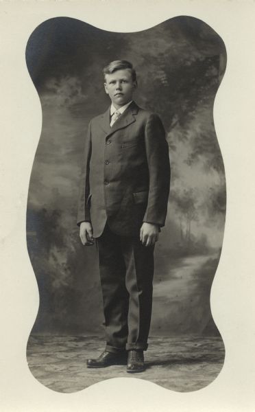 Full-length studio portrait of a young man standing in front of a painted backdrop. A wavy white mask frames the portrait.