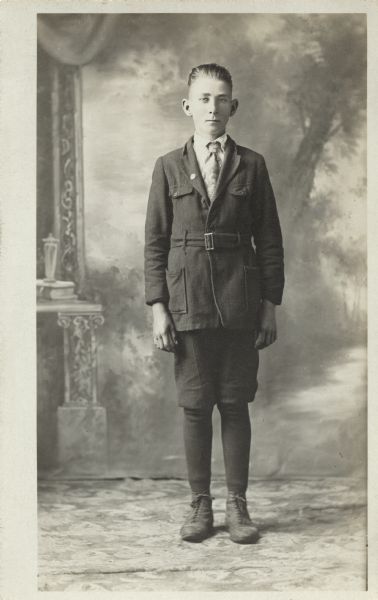Full-length studio portrait of a young man in front of a painted backdrop. He is wearing a suit with short pants, stockings and shoes.