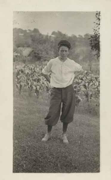 Outdoor portrait of a young man standing in front of a field of corn. His hands are resting behind him on his lower back. He is wearing a cap, shirt and knickers (knickerbockers) with stockings and shoes. The roof of a building can be seen in the far background, with trees and hills in the distance.
