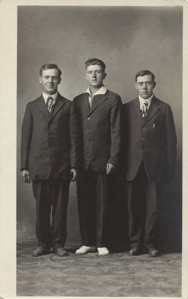 Full-length studio portrait of three men standing in front of a painted backdrop. They are all wearing suits, and the man in the center has white shoes.