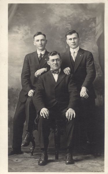 Full-length studio portrait of three men in front of a painted backdrop. Two men, Mel Endres (left) and Math Endres (right), are perched on the chair arms with their hands on the seated (Michael J. Endres) man's shoulders. They are all wearing suits, two with ties and one with a bow tie. All three have pins in their lapels.