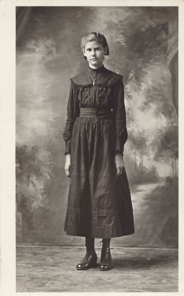 Full-length studio portrait of a young woman standing in front of a painted backdrop. She is wearing a dark dress, hair bow and shoes. A crucifix is hanging on a chain around her neck.