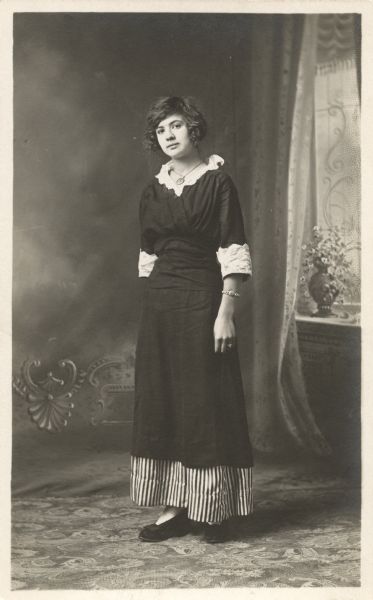 Full-length studio portrait of Anna Bollenbeck Bowar, standing in front a painted backdrop. She is wearing a dark dress with white collar and cuffs, a striped underskirt, necklace, ring and bracelet.