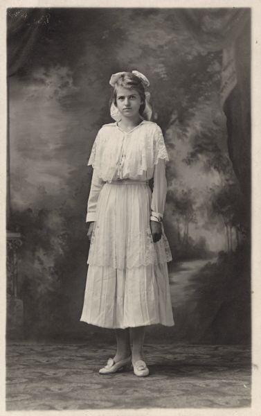 Full-length studio portrait of a young woman standing in front of a painted backdrop. She is wearing a light-colored dress with a lace overlay, hair bow and shoes. A bracelet is on her wrist.