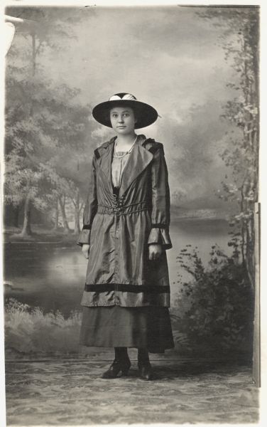Photographic postcard of a full-length studio portrait of a young woman standing in front of a painted backdrop. She is wearing a string of pearls, and a dark dress, hat and shoes.