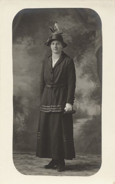 Full-length studio portrait of a woman standing in front off a painted backdrop. She is wearing a dark dress, shoes, hat with bow, light-colored gloves and eyeglasses. In her left hand she holds a small handbag and handkerchief.