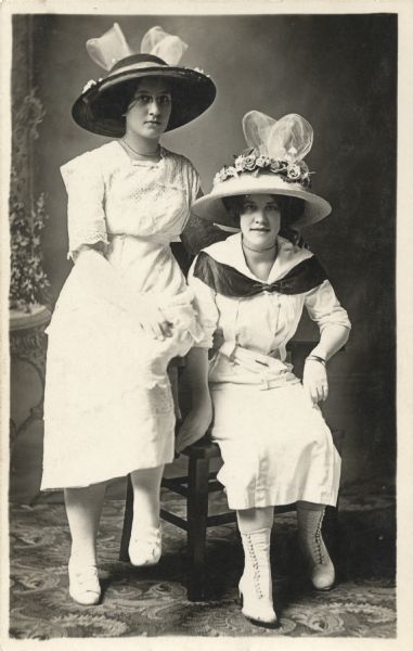 Full-length studio portrait of two women in front of a painted backdrop. One woman is seated, the other is perched on the arm of the chair. They are wearing light-colored dresses, large hats trimmed with flowers and bows, necklaces and a bracelet. One woman wears low shoes, the other, high button shoes. The woman on the left is wearing glasses.