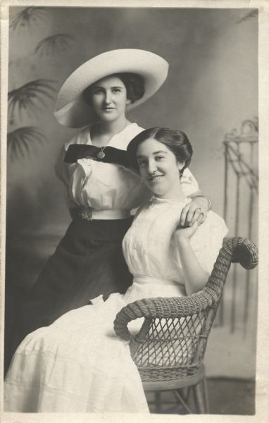 Photographic postcard of a three-quarter length studio portrait of two women in front of a painted backdrop. One woman is seated in a wicker chair, the other is perched on the arm. The seated woman is wearing a light-colored dress, the standing woman is wearing a large hat, light blouse, dark skirt, necklace, bracelet and ring. They are holding hands.