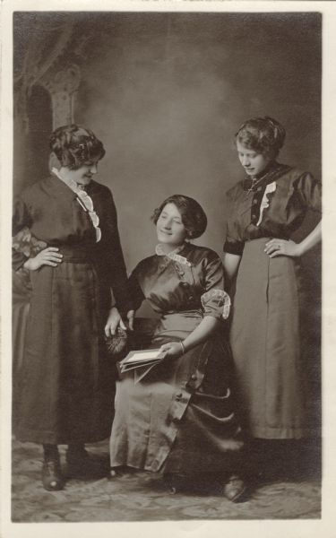 Full-length studio portrait of three women in front of a painted backdrop. One woman is sitting, holding a book; the other two are standing on either side with their hands on their hips, gazing down at her. They are wearing dark dresses with light-colored trim on the bodices. One woman is wearing a necklace, and another woman is wearing a bracelet.