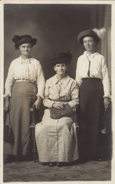 Full-length studio portrait of three women in front of a painted backdrop. One woman is sitting in the center, wearing a light-colored dress and dark hat trimmed with flowers, and holding a handbag. The other two women are standing on either side, wearing dark skirts, light blouses and dark hats trimmed with feathers, also holding handbags. Two of the women are wearing braided necklaces with tassels.