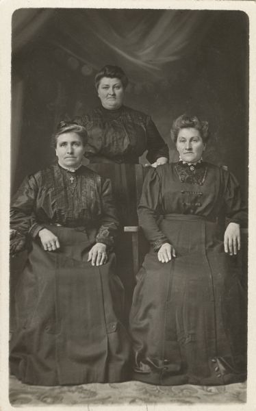 Full-length studio portrait of three women standing in front of a painted backdrop. Two women are seated, and the third woman is standing behind them. All three women are wearing dark dresses. The seated women are wearing jewelry, a pin, crucifix and ring.