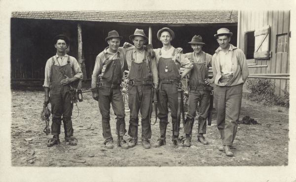 Photographic postcard of an outdoor group portrait of a telephone line crew. The six men are wearing hats and work clothes, and five of the men are wearing belts and straps around their waist, calves and shoes. They appear to be standing in a barnyard, with buildings in the background. Just behind them is a wagon with what looks to be the handles of shovels or other implements hanging over the tailgate.
