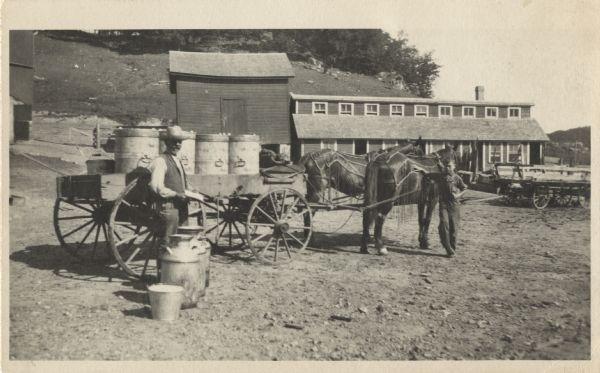Robert Carl Hering and his son Alexander Hering are standing next to a horse-drawn wagon filled with milk containers. Farm buildings are in the background. On the ground near the elder Hering's feet are three containers and a pail. On the right Alexander is standing with two horses wearing fly-nets. In the background is a tree-covered hill.
