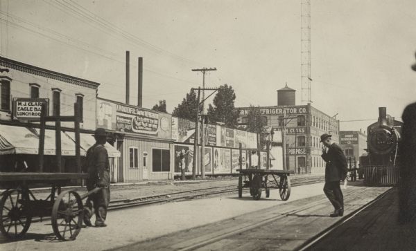 Railroad loading platform with a Gurney Refrigerator Company plant in the far background. On the far left a man pulls a hand cart, in the center a man sits on another cart, and a third man stands smoking on the platform, in front of locomotive 586. Billboards are posted on the wall across the street. Among them, "Fisk," "Old Dutch Cleanser," a refrigerator company, and SpearHead." On the far left is "M.J. Clark, Eagle Bar, Lunch Room."