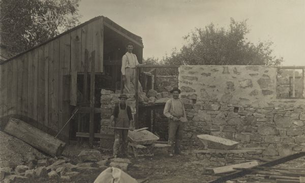 Photographic postcard of three workmen building a stone wall next to a shed.