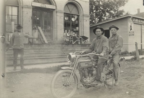 Photographic postcard of two men posing seated on a vintage Harley-Davidson motorcycle. They are dressed in work clothes and hats. Another man with his arm lifted stands on the sidewalk in front of a hardware store. The sign above the door reads, "Tools and Cutlery." Wheelbarrows are on display on the steps and merchandise is in the windows. The building on the right has signs that read, "Valvoline Motor Oil" and "IHC."