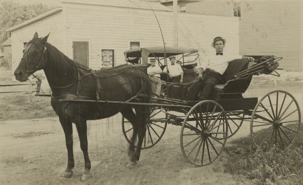 Photographic postcard of a man sitting in a buggy drawn by a horse wearing a fly-net. Behind him is an automobile with two men leaning against the side. All three men are wearing light-colored shirts, dark neckties, and hats. In the background is Matthew Witt's Photography Studio. Samples of the studio's work are on display in a case next to the door. The side of a larger building is on the right.

