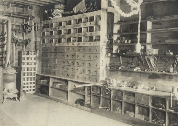 Interior view of a machine shop, possibly for wagon or vehicle repair. A wood burning stove is on the left. In the center is a bank of drawers and cubby holes for tools and parts. On the right are more shelves for tools. Among the tools are metal shears, drills, wrenches, hacksaws, a vise and oil cans. Parts hang on the wall above the stove and drawers. An electric light socket without a bulb hangs down in the right foreground and two more sockets with bulbs hang in the center.