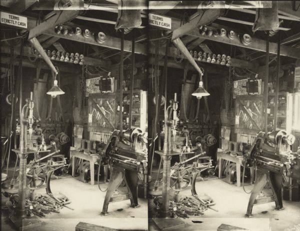 Stereographic print of the interior of a machine shop. The drill press, engine lathe and power hacksaw are powered by a line shaft mounted on the ceiling. Pulleys of many sizes and a bundle of chain are hanging on nails in the ceiling joists. A work light dangles overhead. A sign near the ceiling reads, "Terms Strictly Cash." Some of the tools are calipers, drill bits, etc.