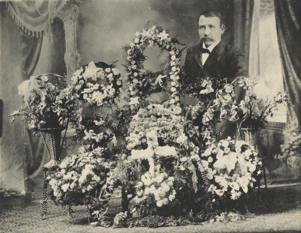 Photographic postcard of a composite studio portrait of funeral floral arrangements and a man in front of a painted backdrop. A head and shoulders portrait of a man was trimmed and attached to the photo and re-photographed.