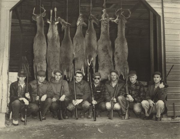 Eight deer hunters pose with seven deer carcasses. The men are squatting in a line inside an open shed door holding their guns. The deer carcasses are hanging in a line from the rafters behind them.