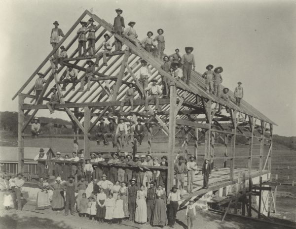 Large group of people posed in, on and around the frame of a barn at a barn raising. A smaller building is in the background on the left. Cultivated fields and wooded hills are in the far background.
