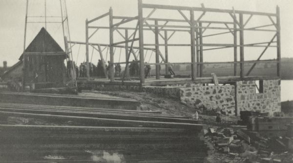 Barn raising, showing the stone foundation and timbers stacked in a pile in the foreground. A smaller building is on the left behind the tower support for a windmill. The workers are on the far side of the barn, raising a section using long poles.