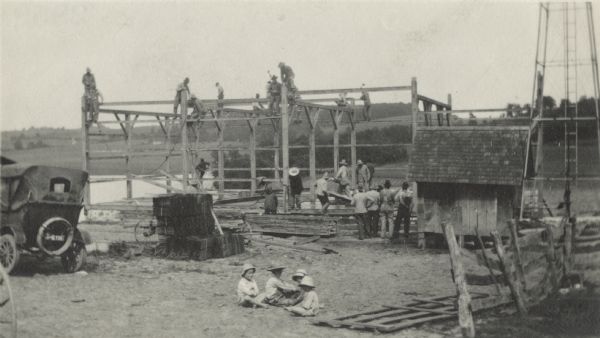 Group of men working on a barn raising, showing some of the workers up on the frame securing the timbers, and other workers carrying more timbers into position. A group of four small children sit on the ground in the foreground. A smaller building is on the right beside the tower support for a windmill. The rear of an automobile and a pile of bound shingles are on the left. In the background are wooded hills.